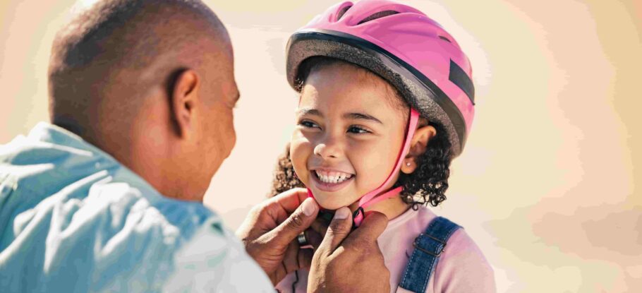 father putting a pink helmet on young daughter before she gets on bicycle