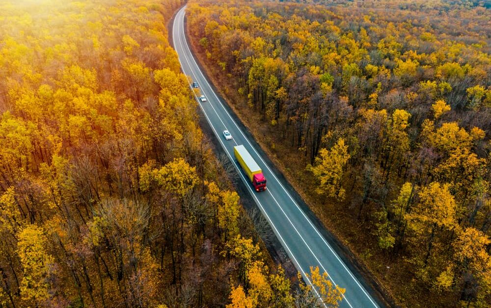 fall scene with orange and red leaves, highway in the middle of woods with a tractor-trailer and three cars