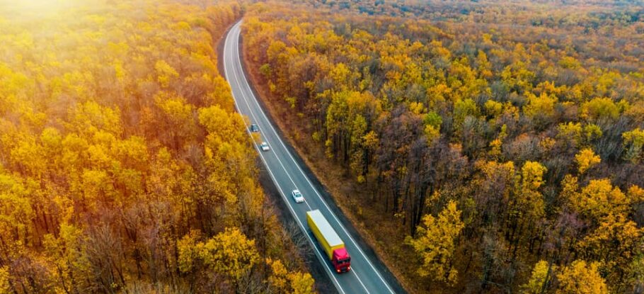 fall scene with orange and red leaves, highway in the middle of woods with a tractor-trailer and three cars